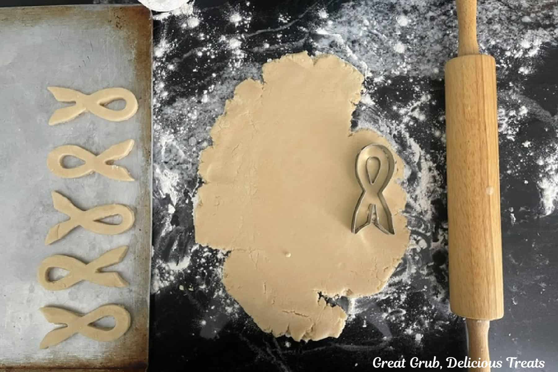 A black granite countertop with sugar cookie dough rolled out with a rolling pin and a ribbon cookie cutter placed on the dough, and cut out cookies on a baking sheet.