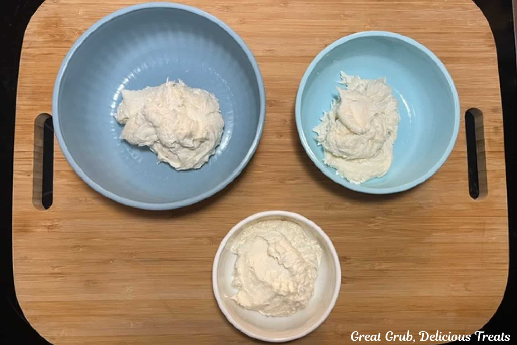 A wood cutting board with three bowls filled with white buttercream frosting.
