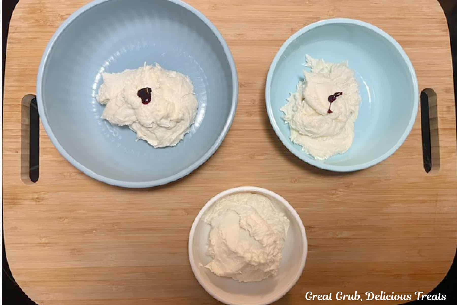 Three bowls on a wood cutting board filled with buttercream frosting with food coloring drops added to two of the bowls of frosting before being mixed together.