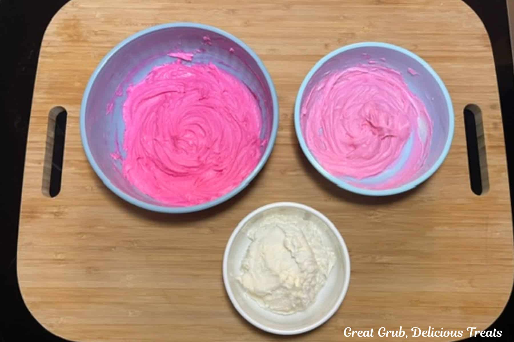 A wood cutting board with three bowls. One with white, one with light pink, and one with bright pink buttercream frosting.