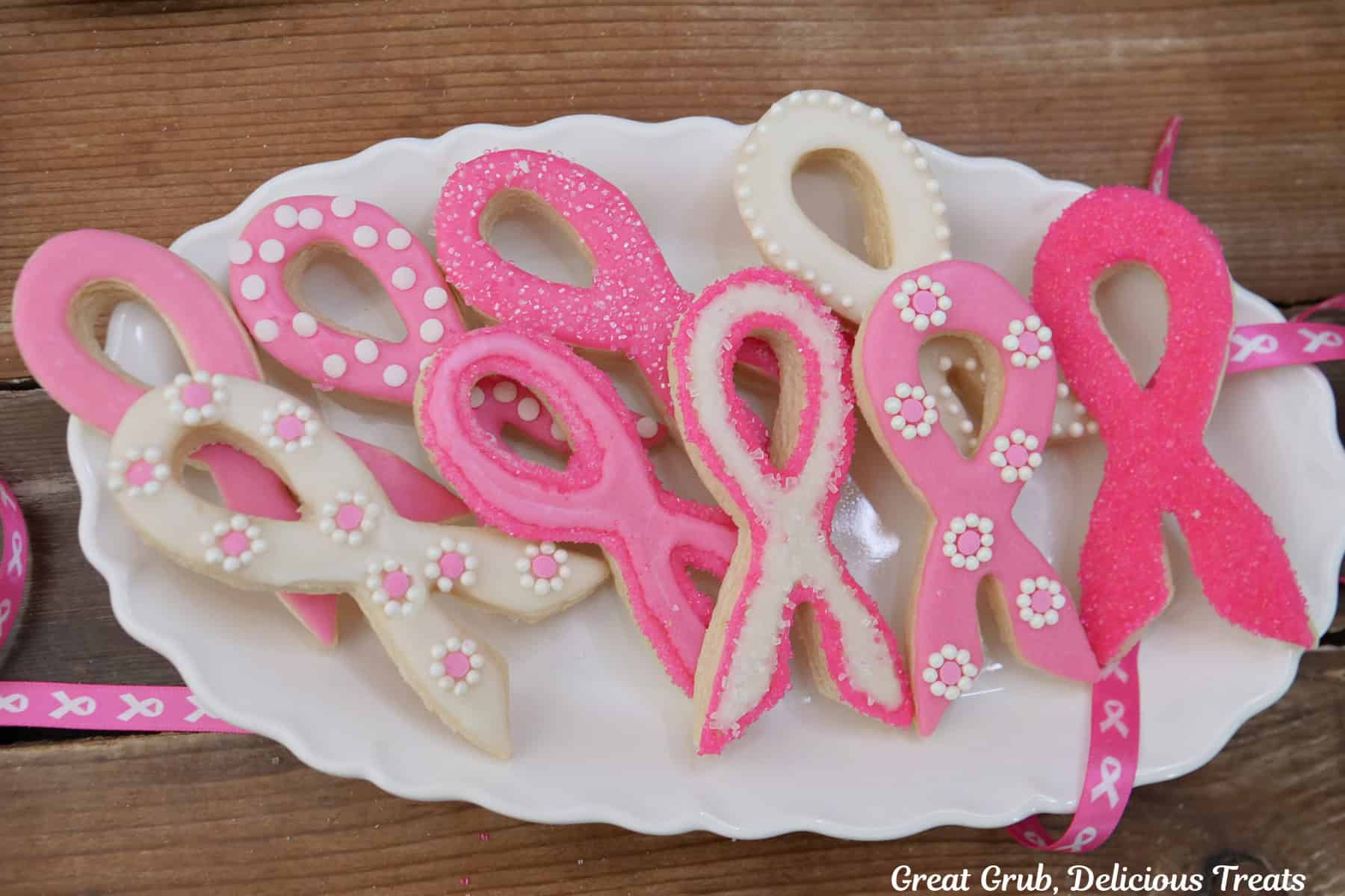 A wood surface with a white oval plate with eight breast cancer ribbon cookies on it decorated with white and pink icing and candy sprinkles.