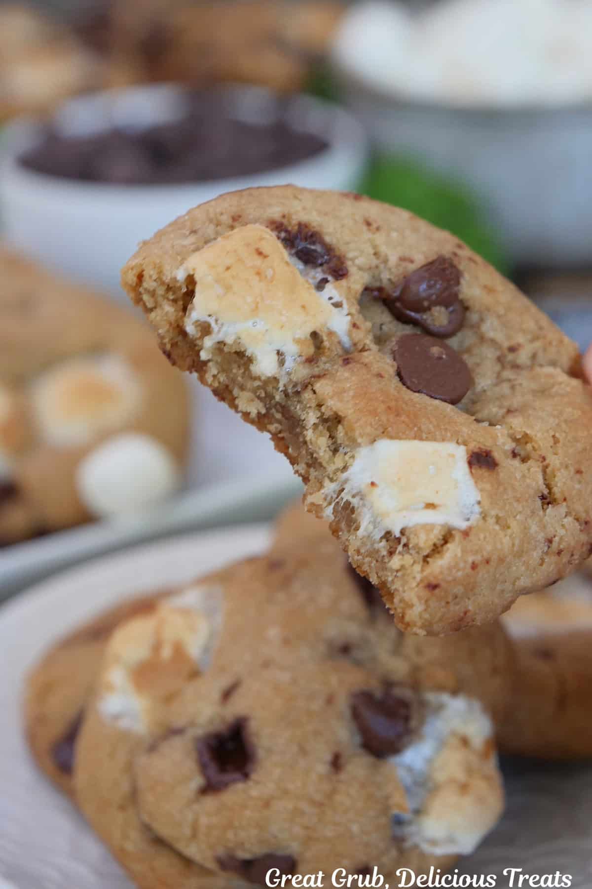 A brown butter s'mores cookie with a bite taken out of it held above more cookies on a plate below.