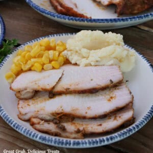 A white plate with blue trim with three slices of smoked Cajun turkey breast on it with corn and mashed potatoes.