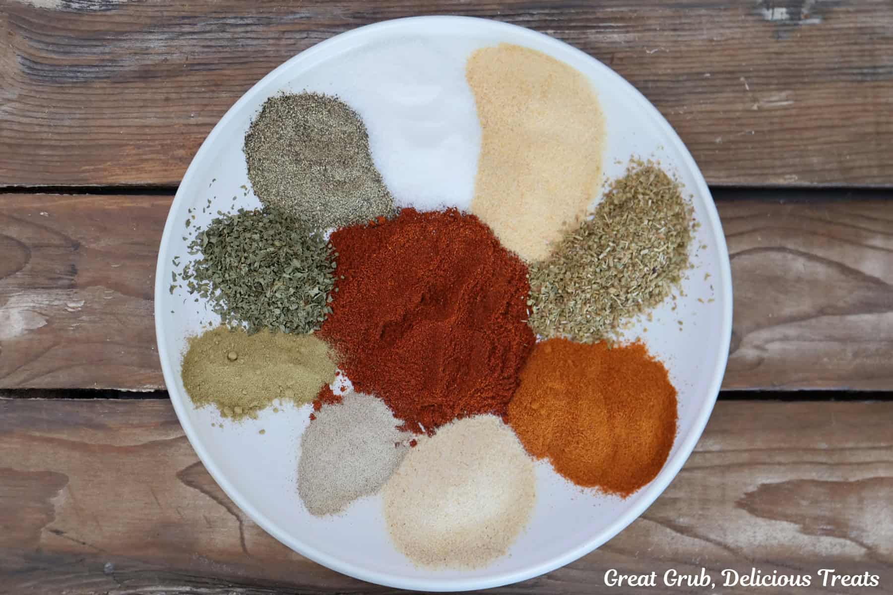 A wood surface with a round white shallow bowl with ten different seasonings on it.