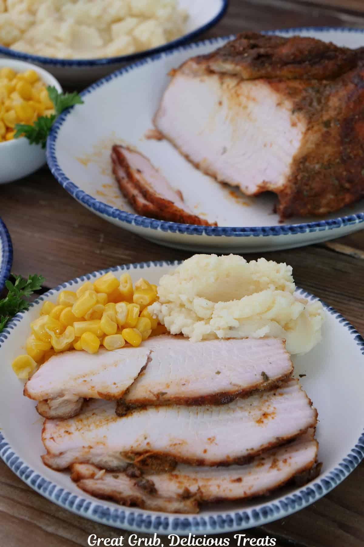 A white plate with blue trim with three slices of Cajun smoked turkey breast on it with mashed potatoes and corn, and the smoked turkey breast roast in the background.