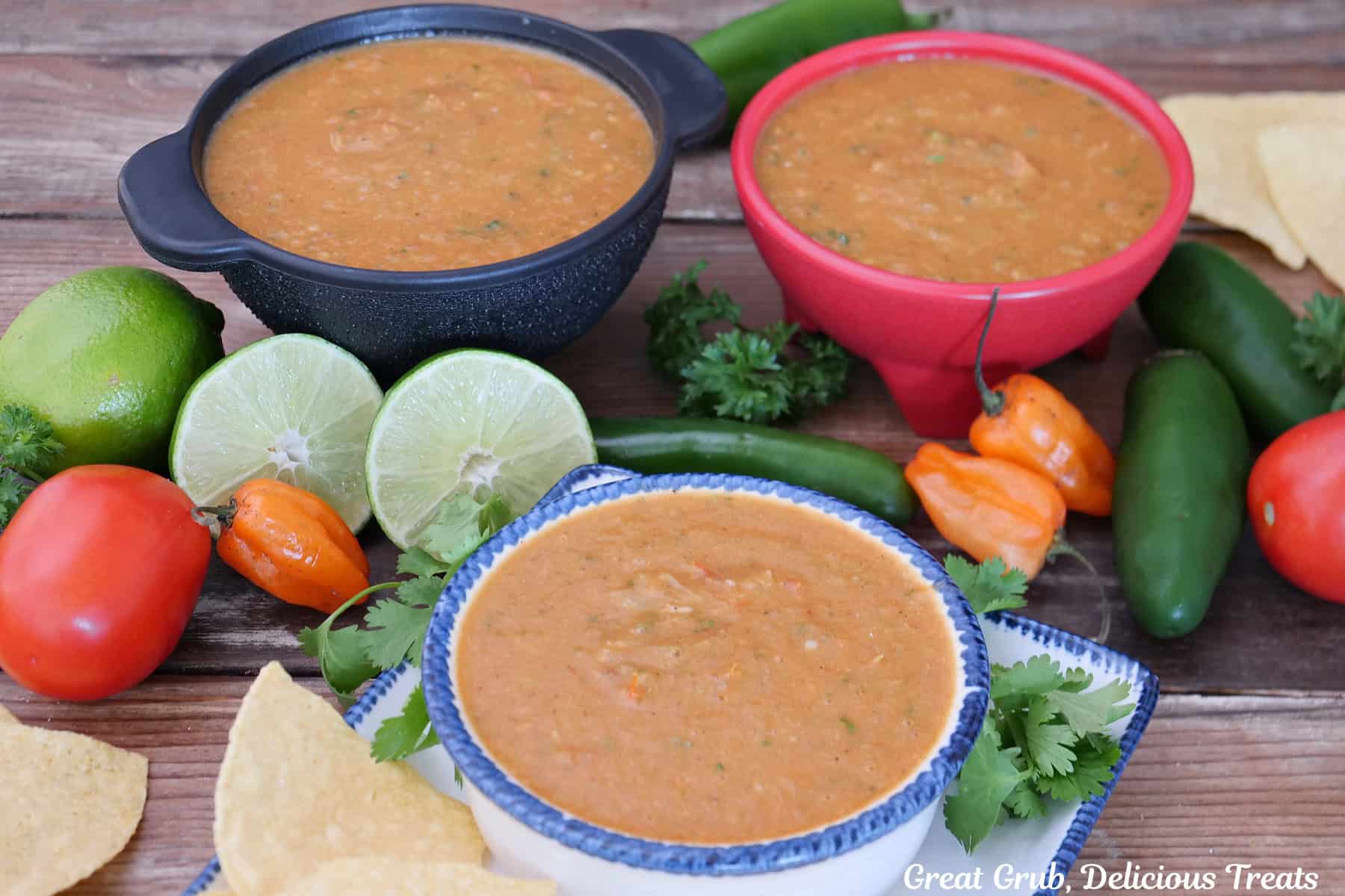 Three bowls with habanero salsa in them with three types of peppers, tomatoes, and onions placed around the bowls along with tortilla chips.