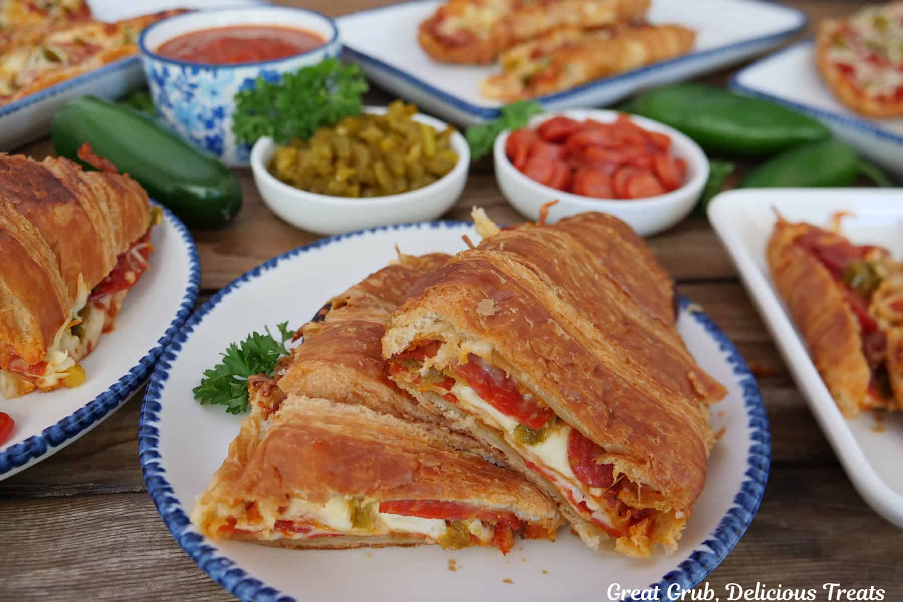 White plates with blue trim with pepperoni pizza croissant sandwiches on them with two small white bowls filled with diced jalapenos and mini pepperoni slices.