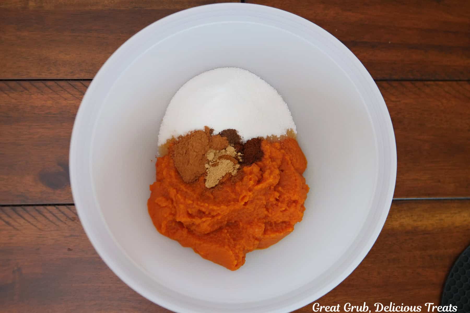 A wood surface with a white mixing bowl filled with pumpkin filling ingredients.