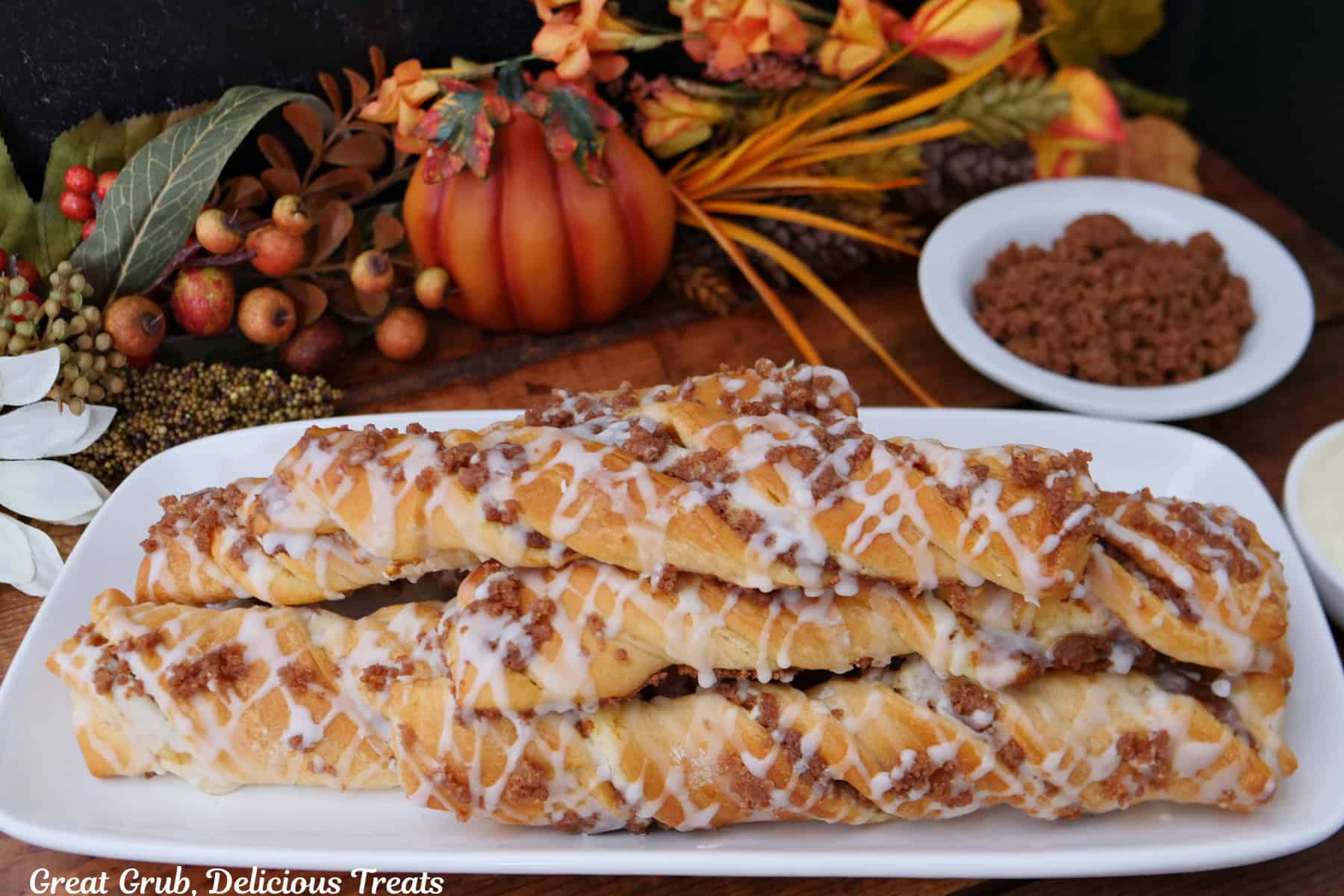 A white oblong plate with six pumpkin cream cheese streusel twists on it with fall foliage in the background.