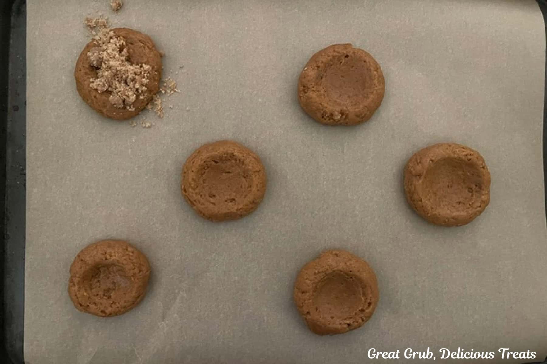 A parchment paper lined baking sheet with six pumpkin cookies on it with the center press in creating a divot.