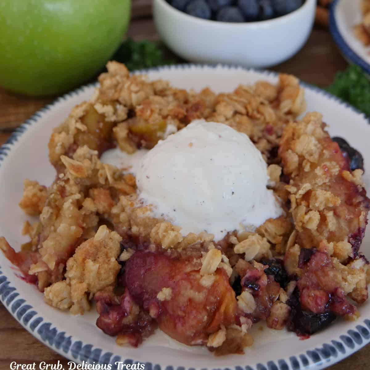 A white plate with blue trim with a serving of sheet pan apple blueberry crisp topped with a scoop of vanilla ice cream on it.