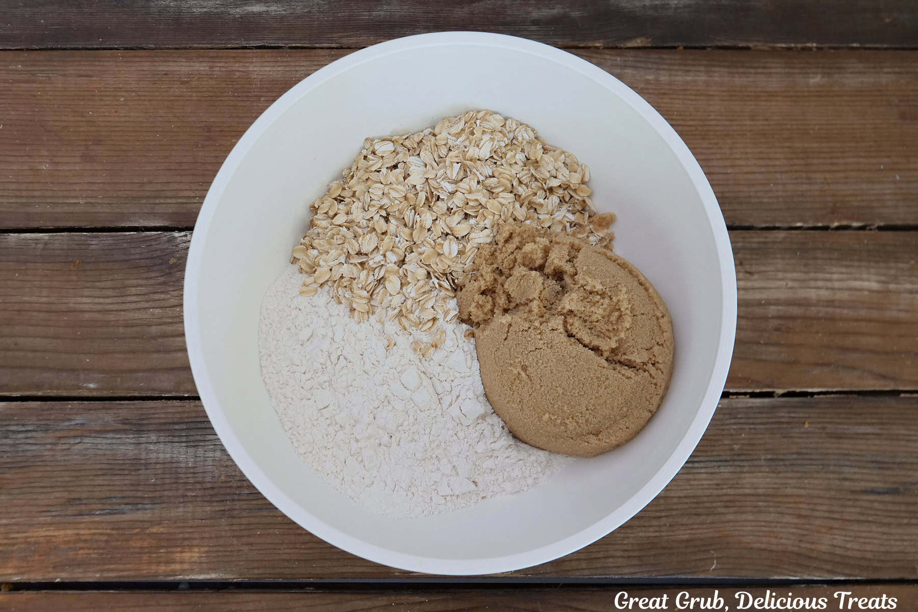 A white mixing bowl with brown sugar, oatmeal, and flour in it before being mixed together.