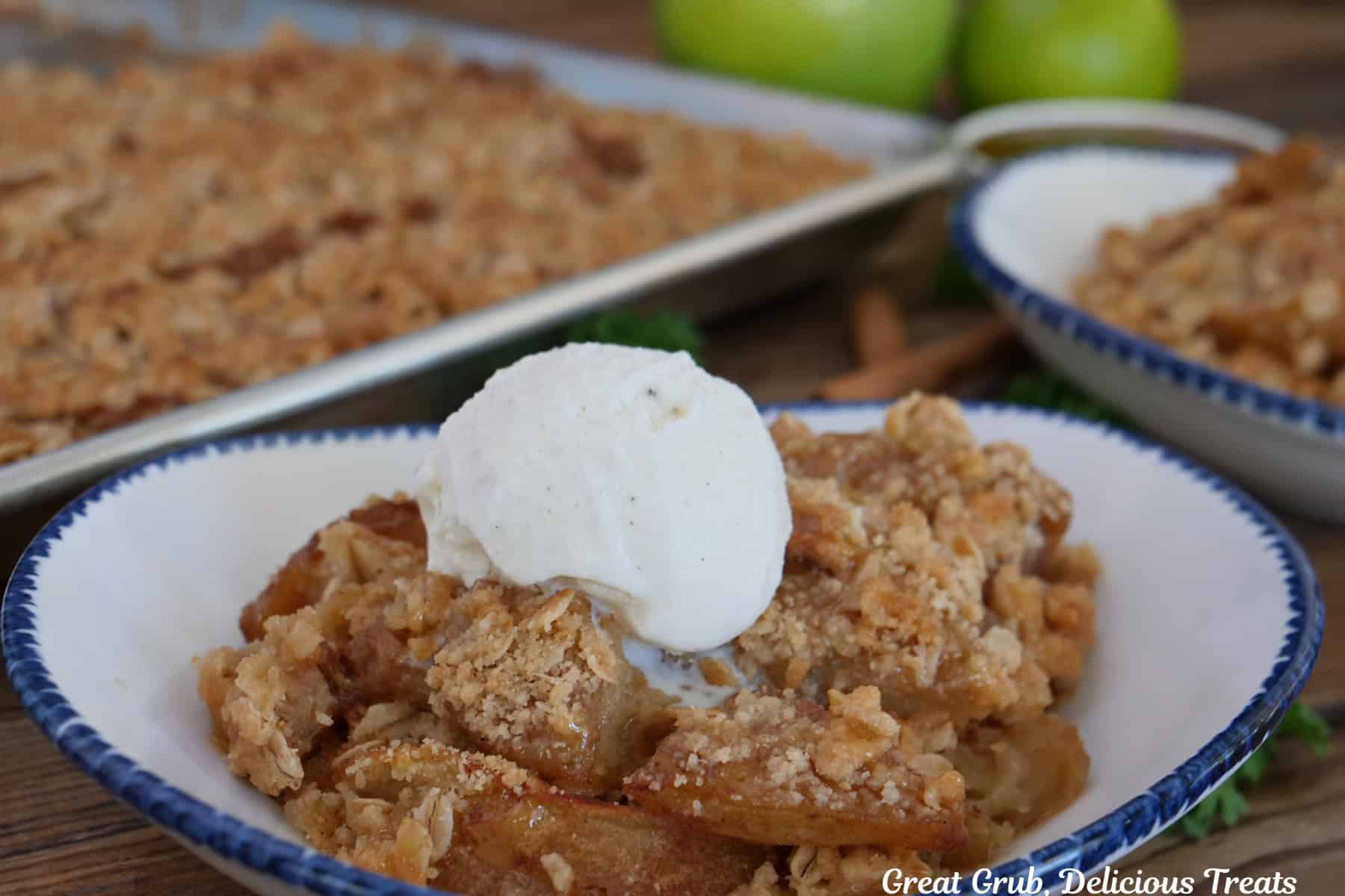 A white bowl with blue trim filled with caramel apple crisp and vanilla ice cream op top, and the sheet pan and apples in the background.