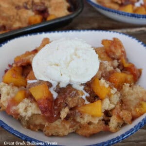 A white bowl with blue trim with a serving of sheet pan peach cobbler in it with a scoop of vanilla ice cream on top.