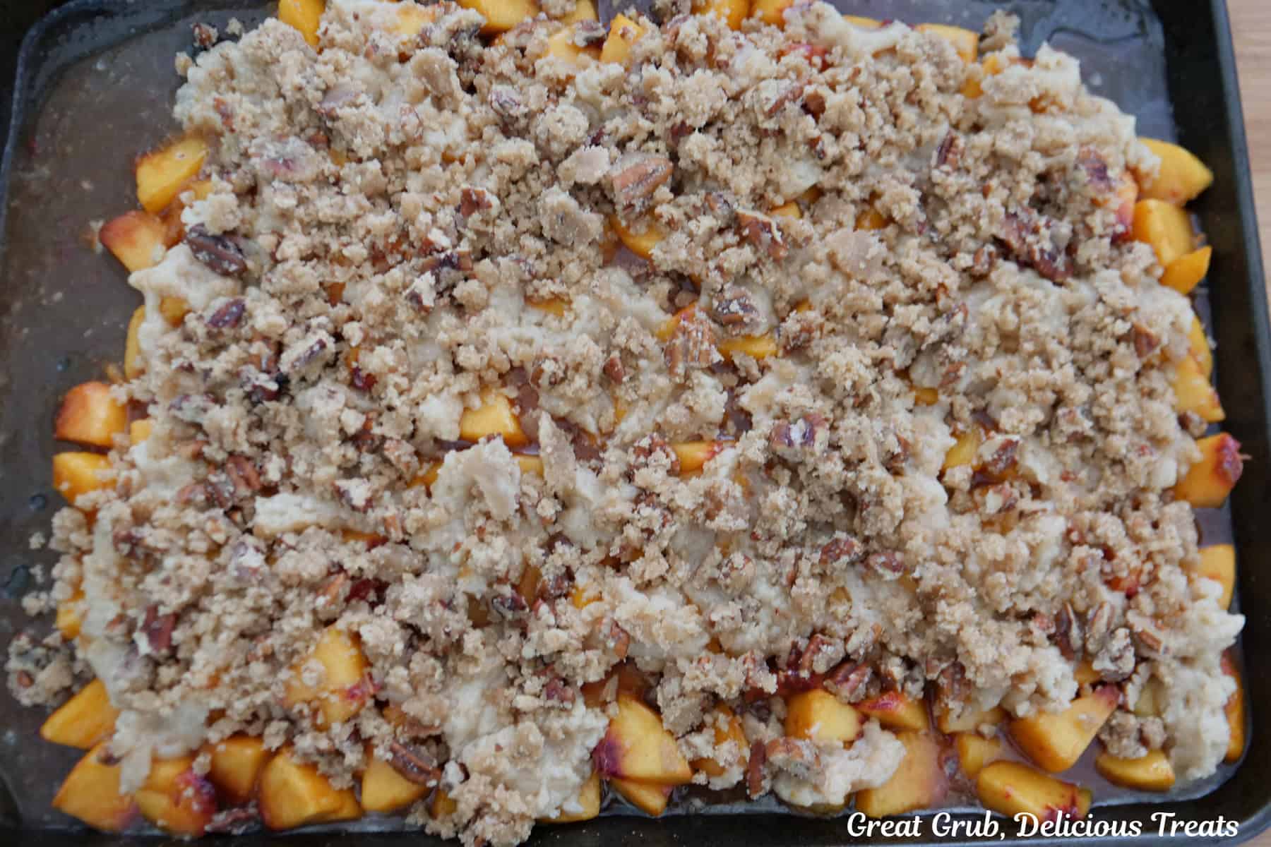A baking sheet with peach cobbler filling, topping, and pecan topping spread out before being baked in the oven.