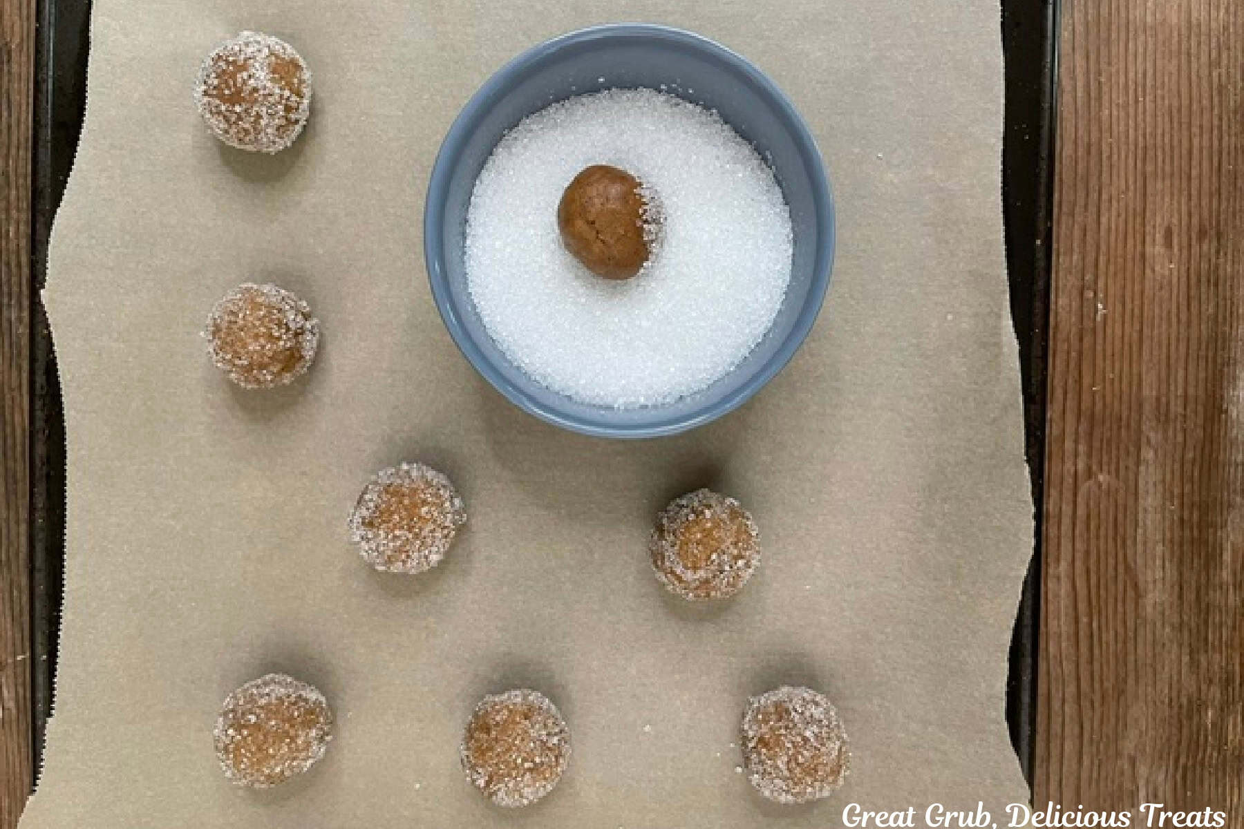 A blue bowl filled with coarse sugar and a molasses cookie ball being rolled into it, with more sugar coated dough balls along side.