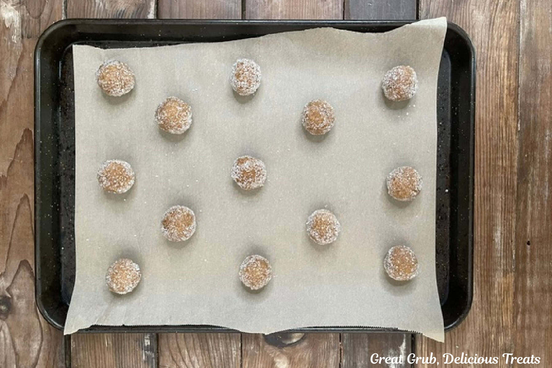 Thirteen molasses cookie dough balls covered in sugar are placed on a parchment line baking sheet before being baked.