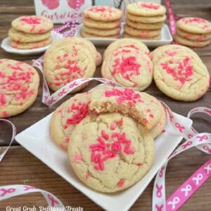 A white plate with four breast cancer sugar cookies on it with more cookies in the background alone with thin pink and white ribbon randomly placed about.