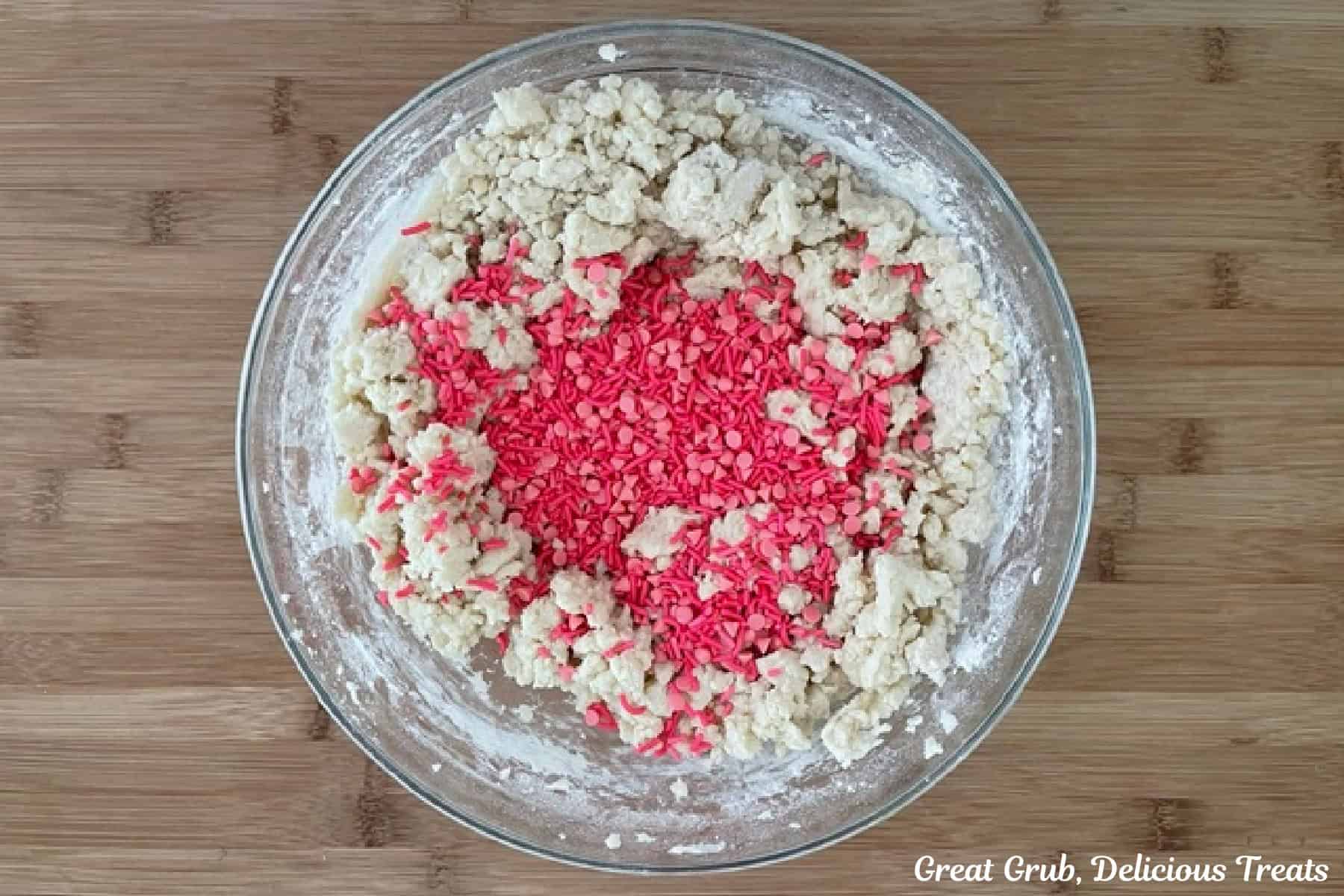 A wood surface with a glass bowl filled with sugar cookie dough and bright pink candy sprinkles added before being mixed together.