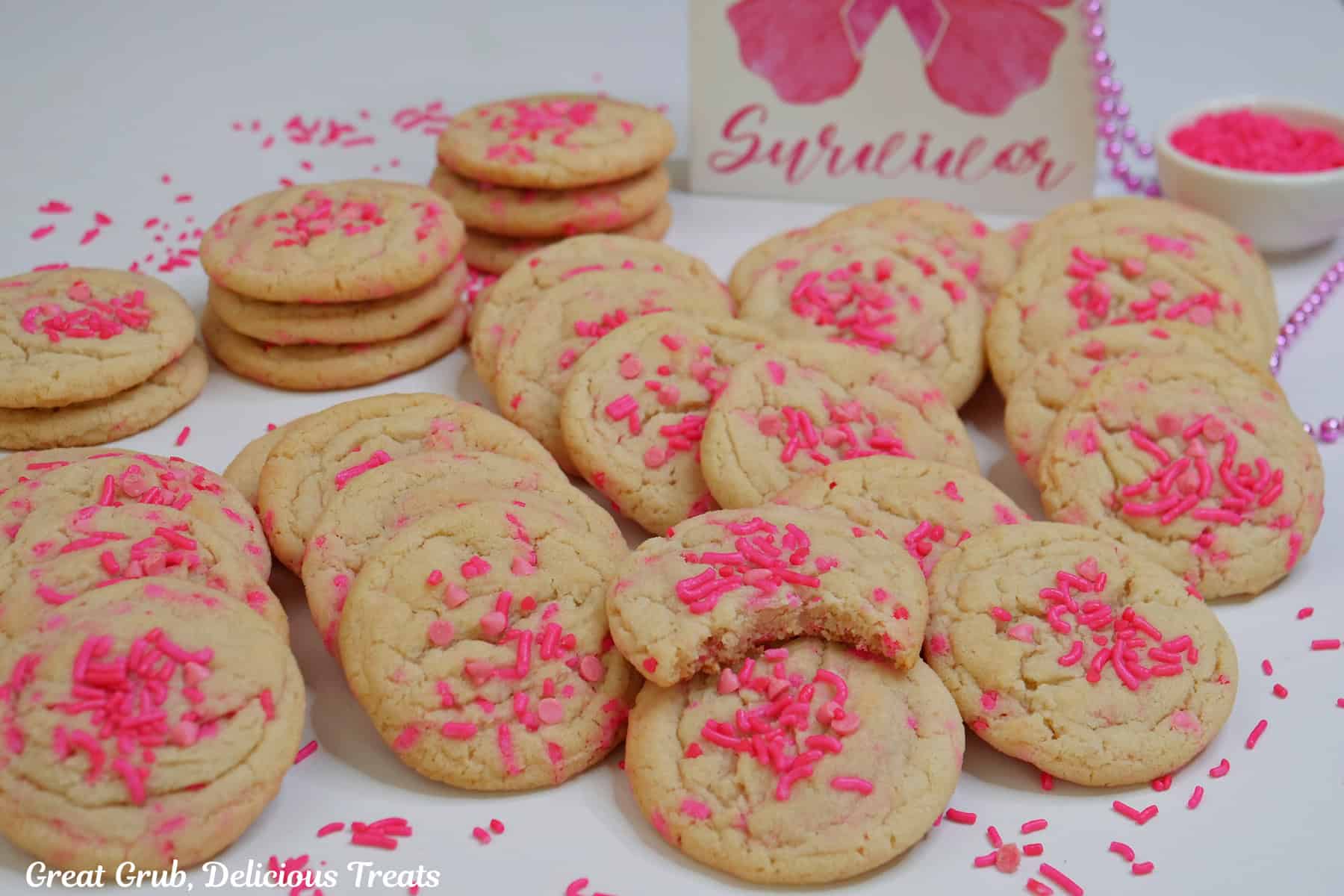 A white surface with a couple dozen breast cancer sugar cookies placed all over the surface with pink candy sprinkles scattered about.