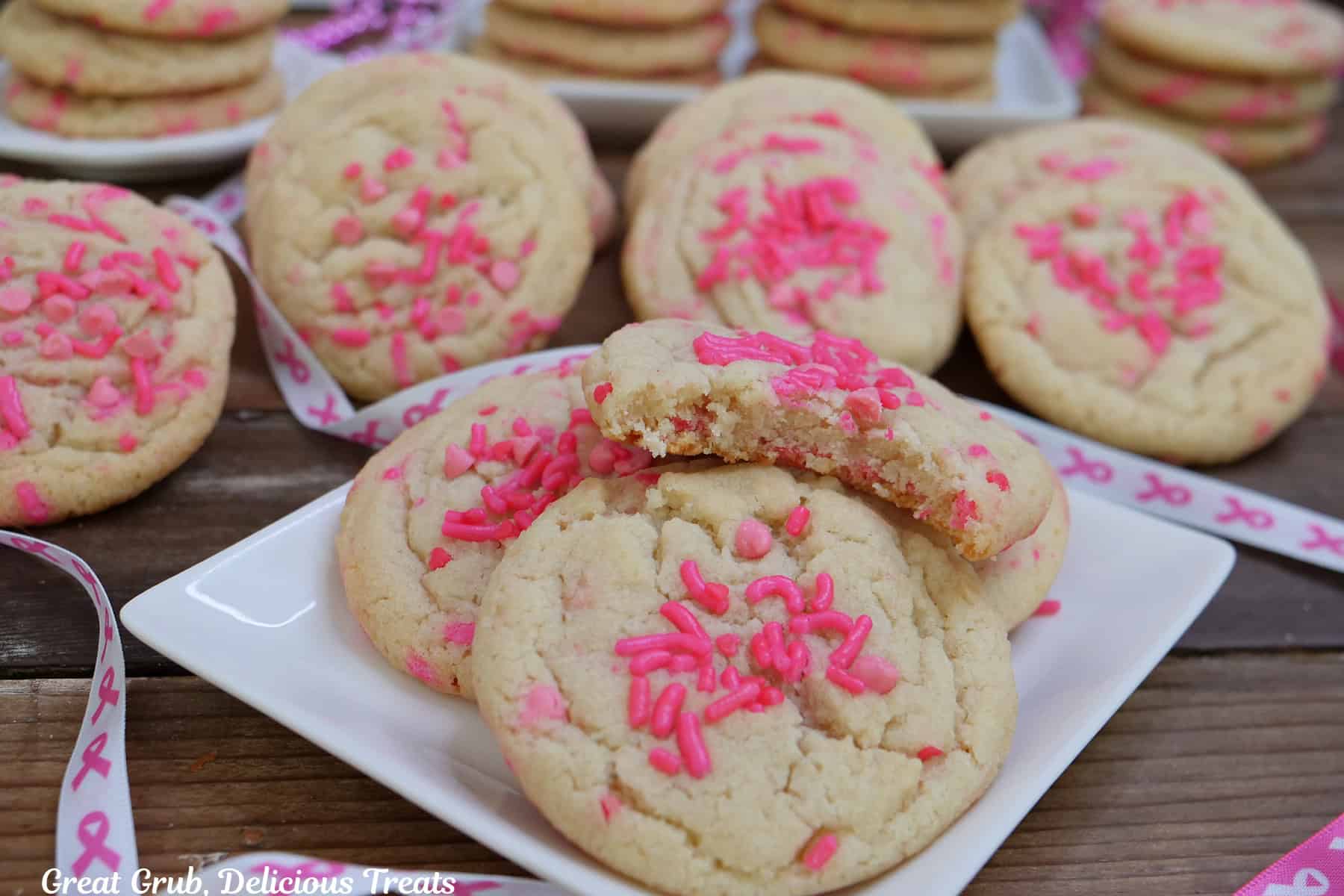 A white plate with four sugar cookies on it with a bite taken out of the one on top, with more cookies in the background.