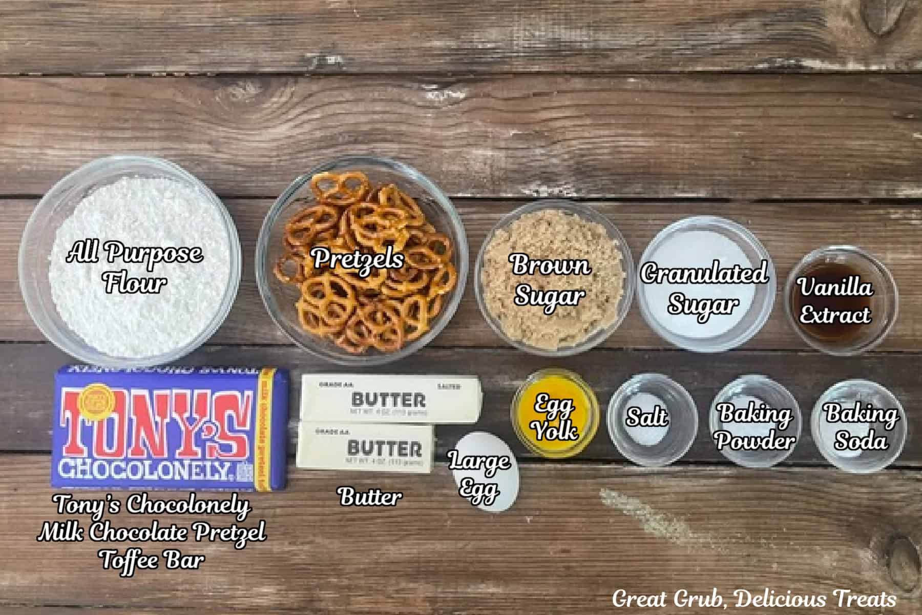 A rustic wood surface with all the ingredients placed on it to make brown butter pretzel toffee cookies.