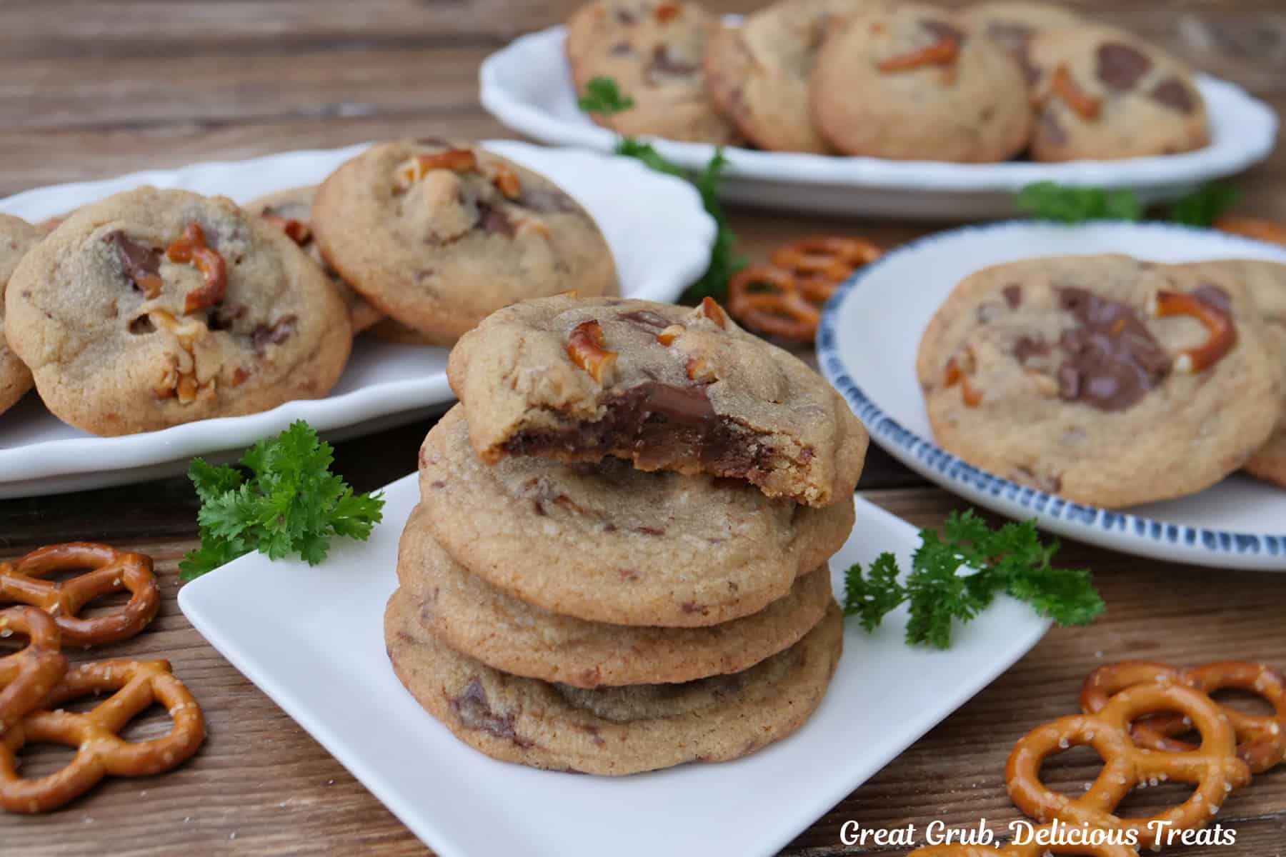 A wood surface with four white plates with brown butter pretzel toffee cookies on them, with pretzels randomly placed about.