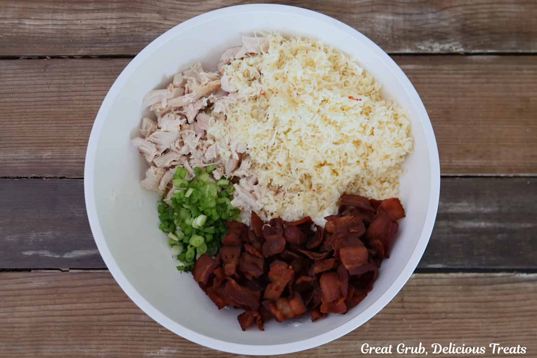 A wood surface with a white bowl filled with chopped rotisserie chicken, bacon, shredded cheese, and green onions.