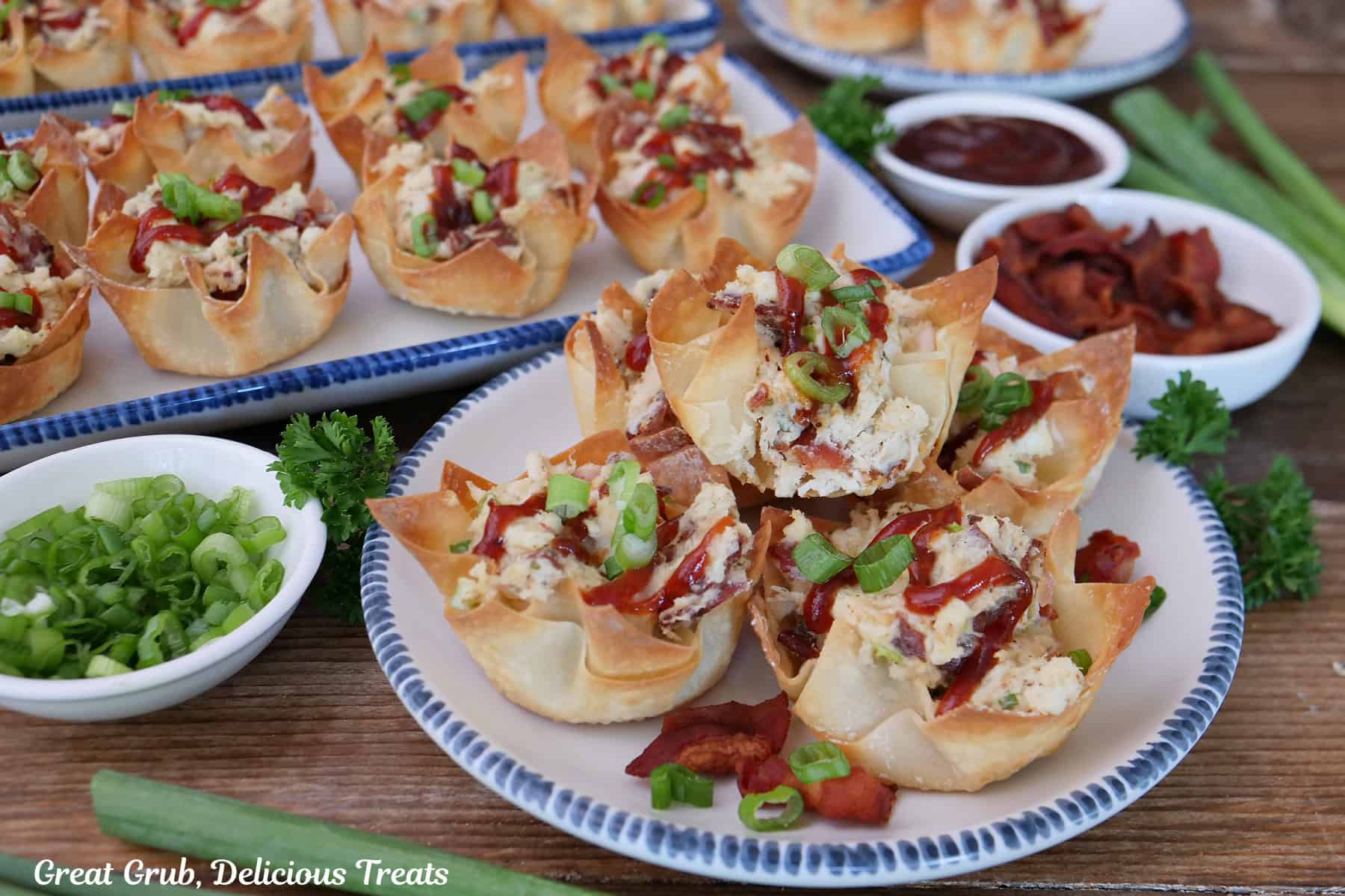 A white plate with blue trim with five chicken bacon ranch wonton cups on it with a bite taken out of the one on top, and more wonton cups in the background.