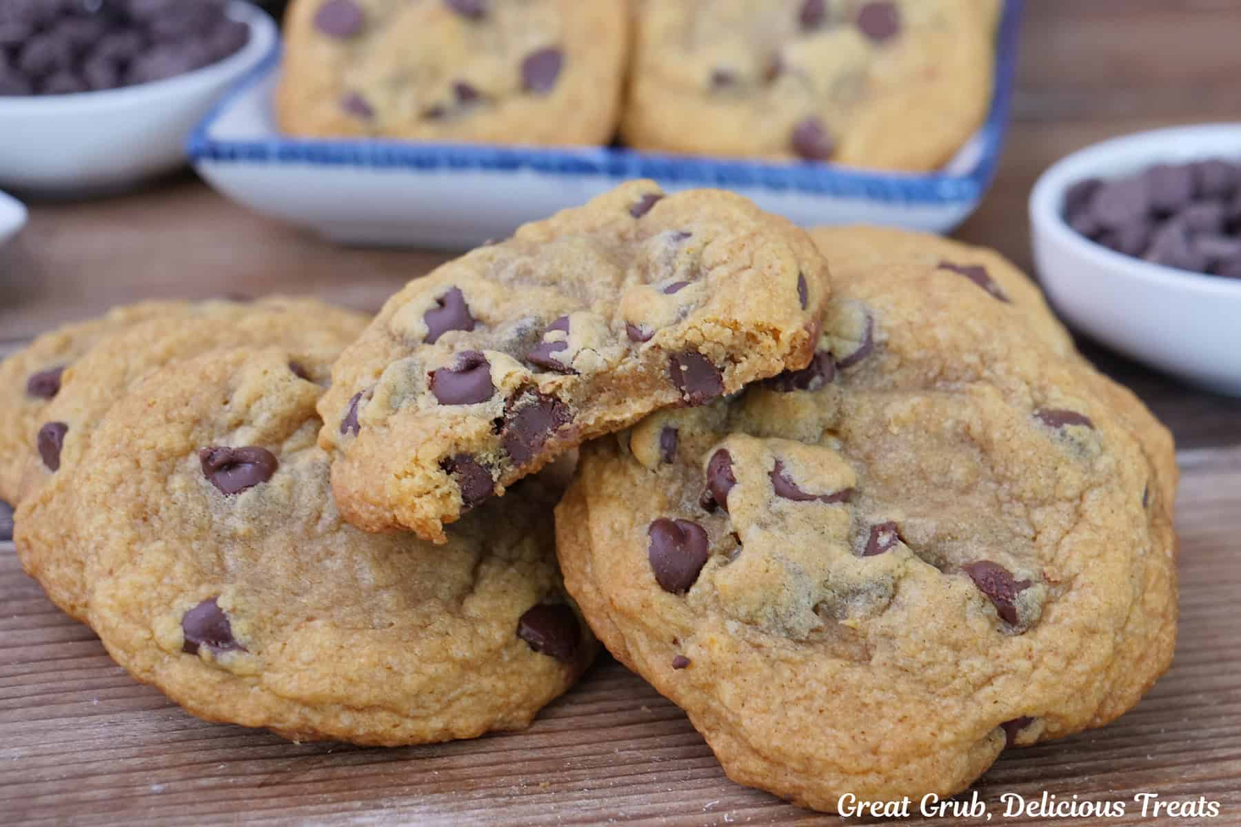 A wood surface with six cookies placed on it with a bite taken out of the cookie on top, with more cookies in the background.