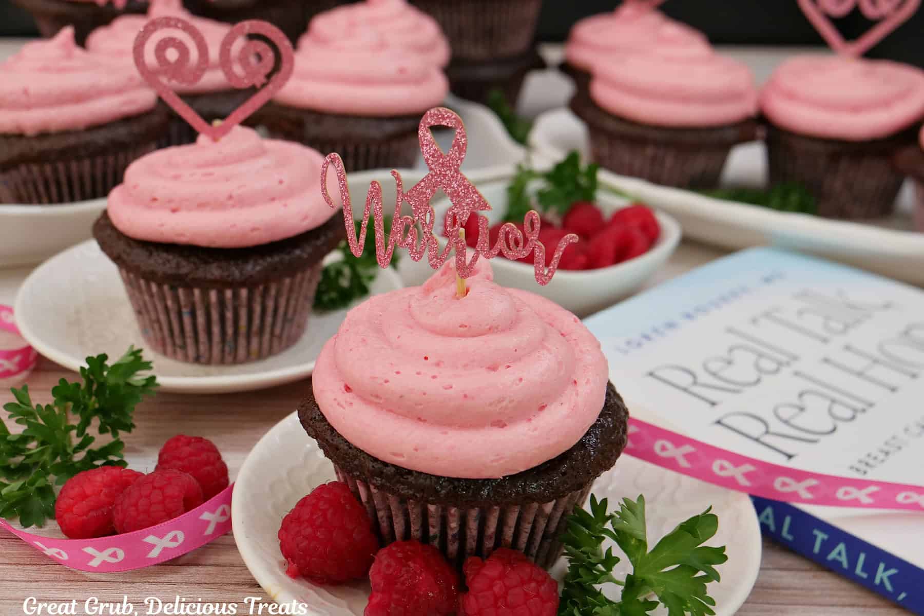 A wood surface with five small white plates with chocolate cupcakes with raspberry buttercream frosting on top, with Dr. Loren Rourke's Real Talk, Real Hope off to the side, and fresh raspberries randomly placed about.