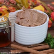 A white bowl filled with homemade cinnamon honey butter with a small decorative bottle of honey off to the side, and cinnamon sticks placed in front of the bowl, and fall foliage in the background.