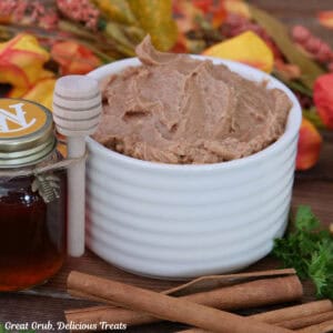 A white bowl filled with homemade cinnamon honey butter with a small decorative bottle of honey off to the side, and cinnamon sticks placed in front of the bowl, and fall foliage in the background.
