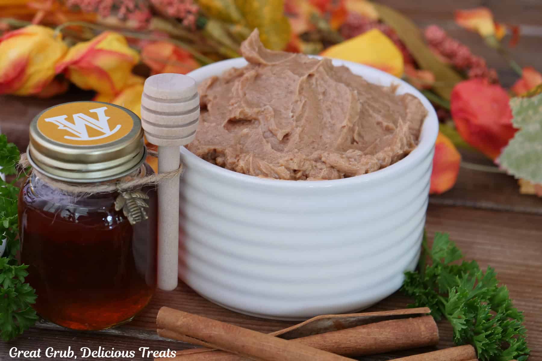 A wood surface with a white bowl filled with cinnamon honey butter, a small decorative bottle of honey on the left side, and fall foliage in the background.