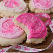 Four pink and white frosted sugar cookies with a bite taken out of the cookie in front, and pink and white ribbon with pink breast cancer awareness ribbons on it.