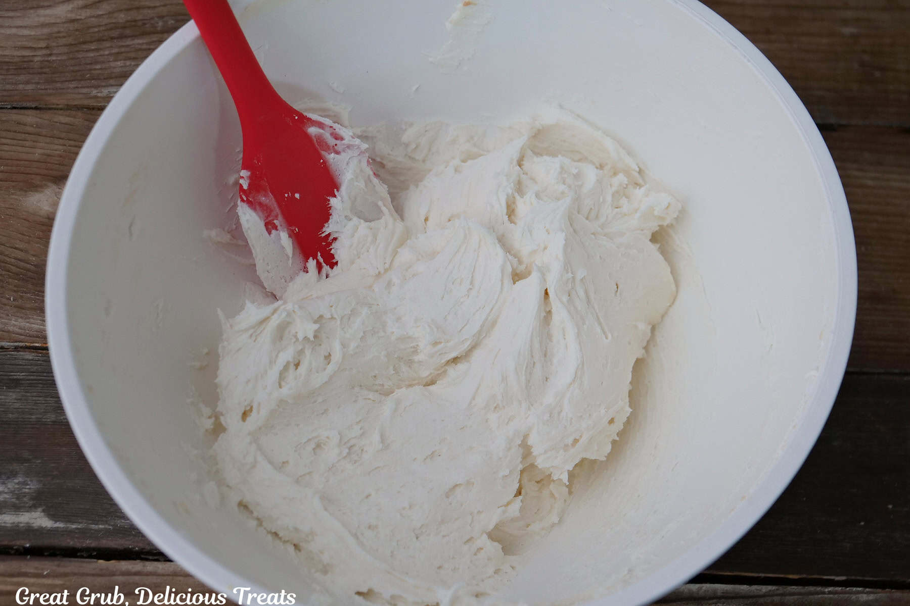 A white mixing bowl with buttercream frosting in it with a red spatula in the bowl.