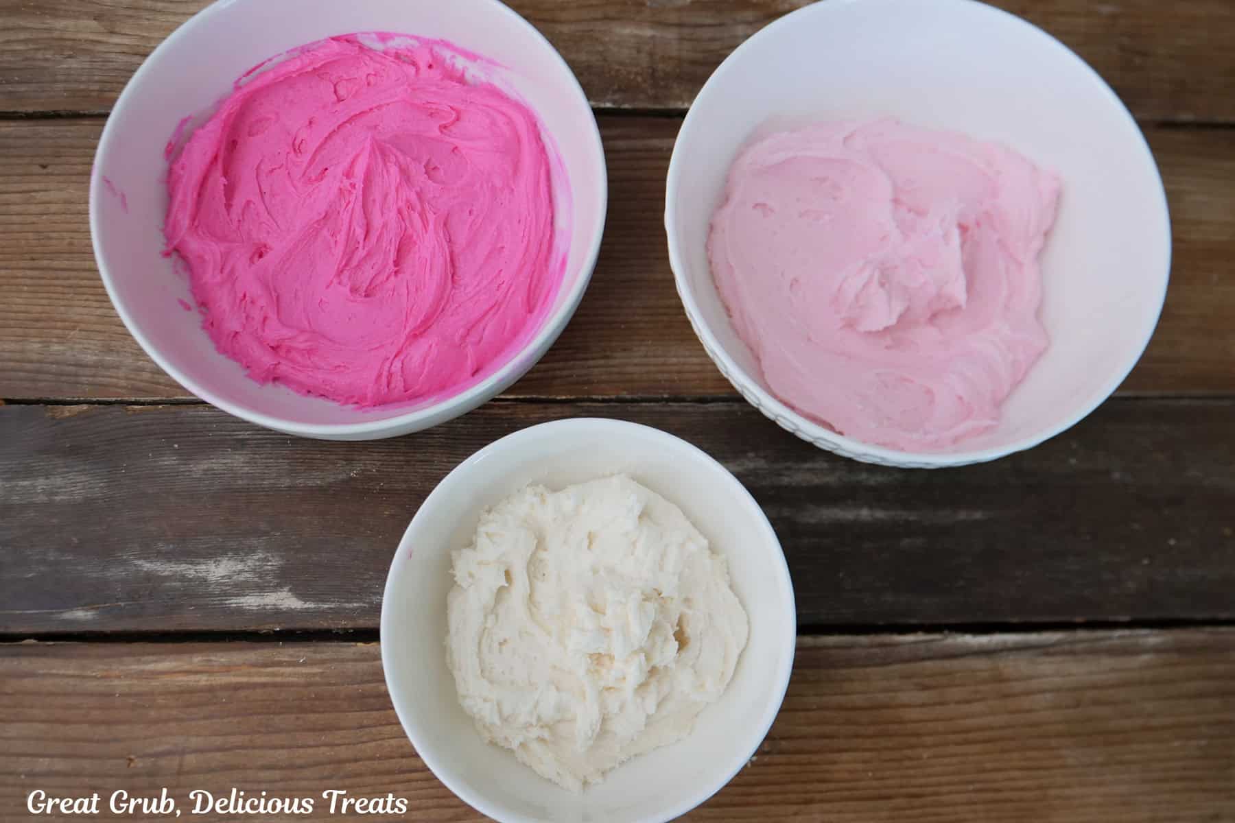 A rustic wood surface with three bowls filled with buttercream, one with white, one with light pink, and one with bright pink frosting.
