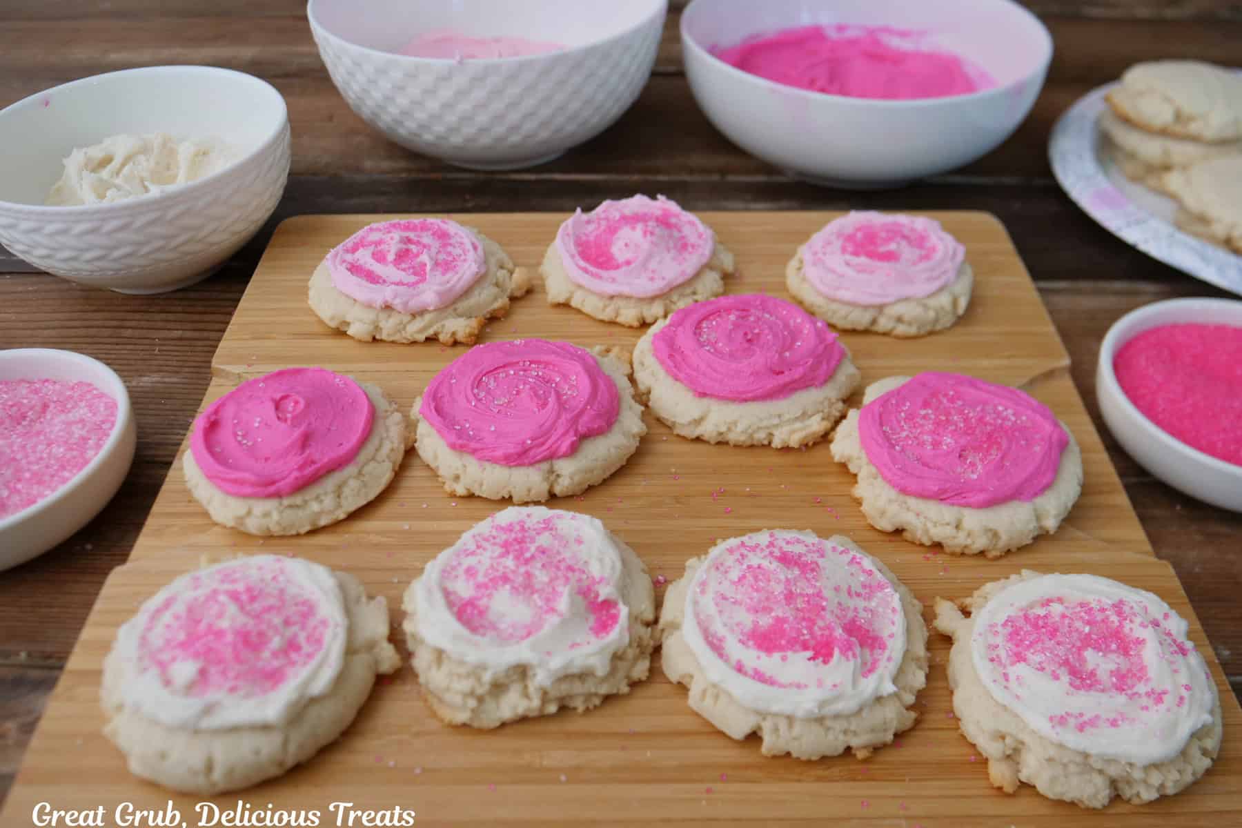 A wood surface with eleven sugar cookies on it frosted with white, pink, and bright pink buttercream frosting and candy sprinkles, and white bowls filled with the three colors of frosting, and two with candy sprinkles in them.