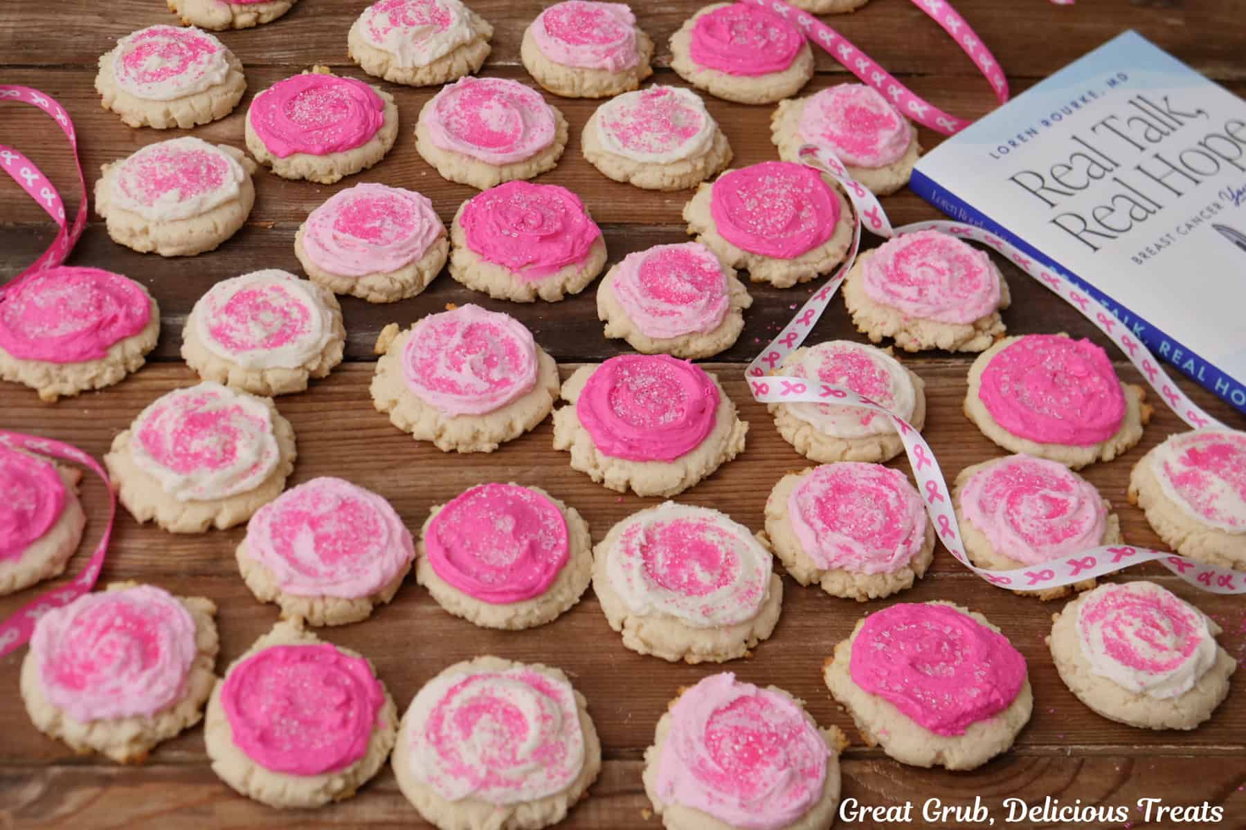 A wood surface with 3 dozen frosted sugar cookies with pink and white buttercream, and Dr. Loren Rourke's "Real Talk, Real Hope" book off to the right side.