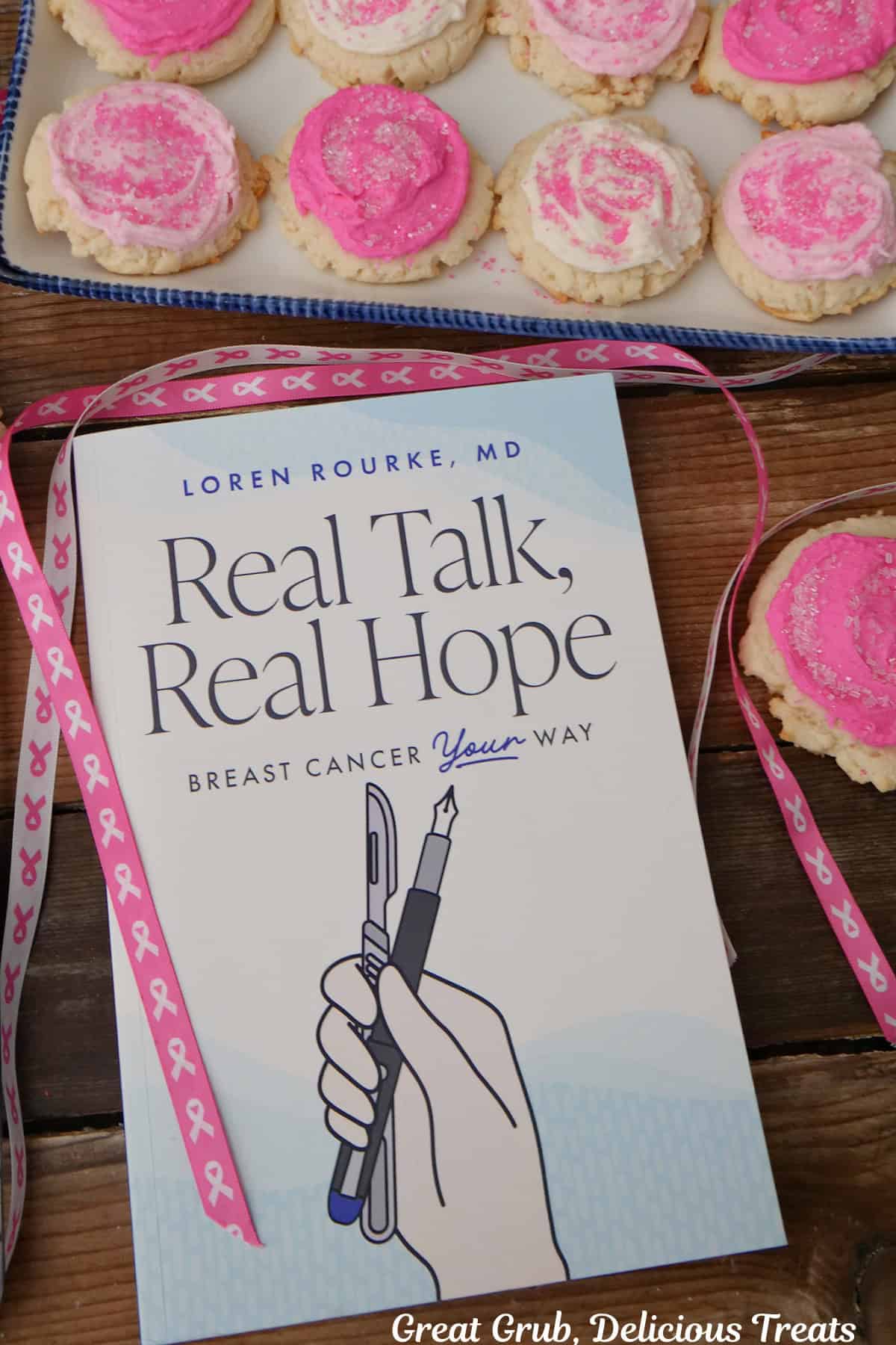 A wood surface with Dr. Loren Rourke's book "Real Talk, Real Hope" placed on it with pink and white decorated sugar cookies and pink ribbon.