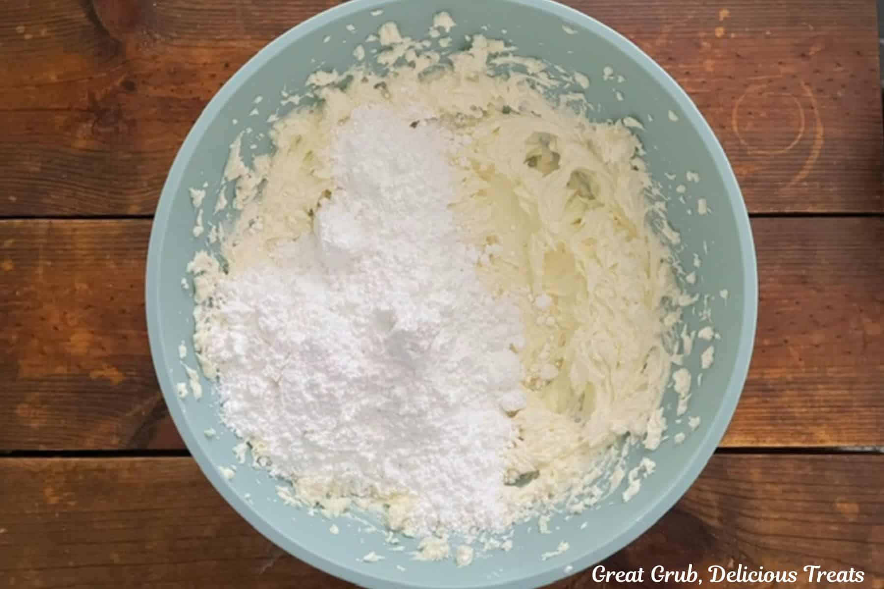 A wood surface with a light blue bowl with powdered sugar added to the cream cheese mixture.