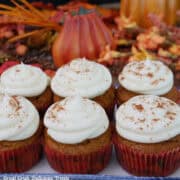 Six pumpkin cupcakes with maple frosting on a white plate with fall foliage in the background.