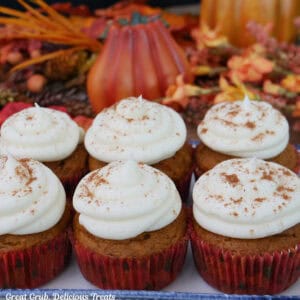 Six pumpkin cupcakes with maple frosting on a white plate with fall foliage in the background.