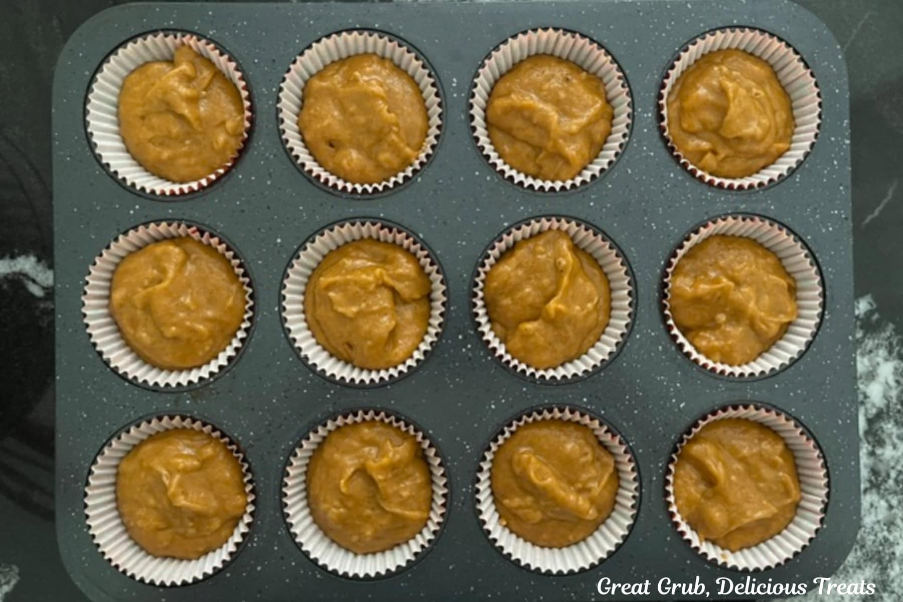 A twelve count cupcake tin with pumpkin batter in each paper liner filled ⅔ full before being baked.