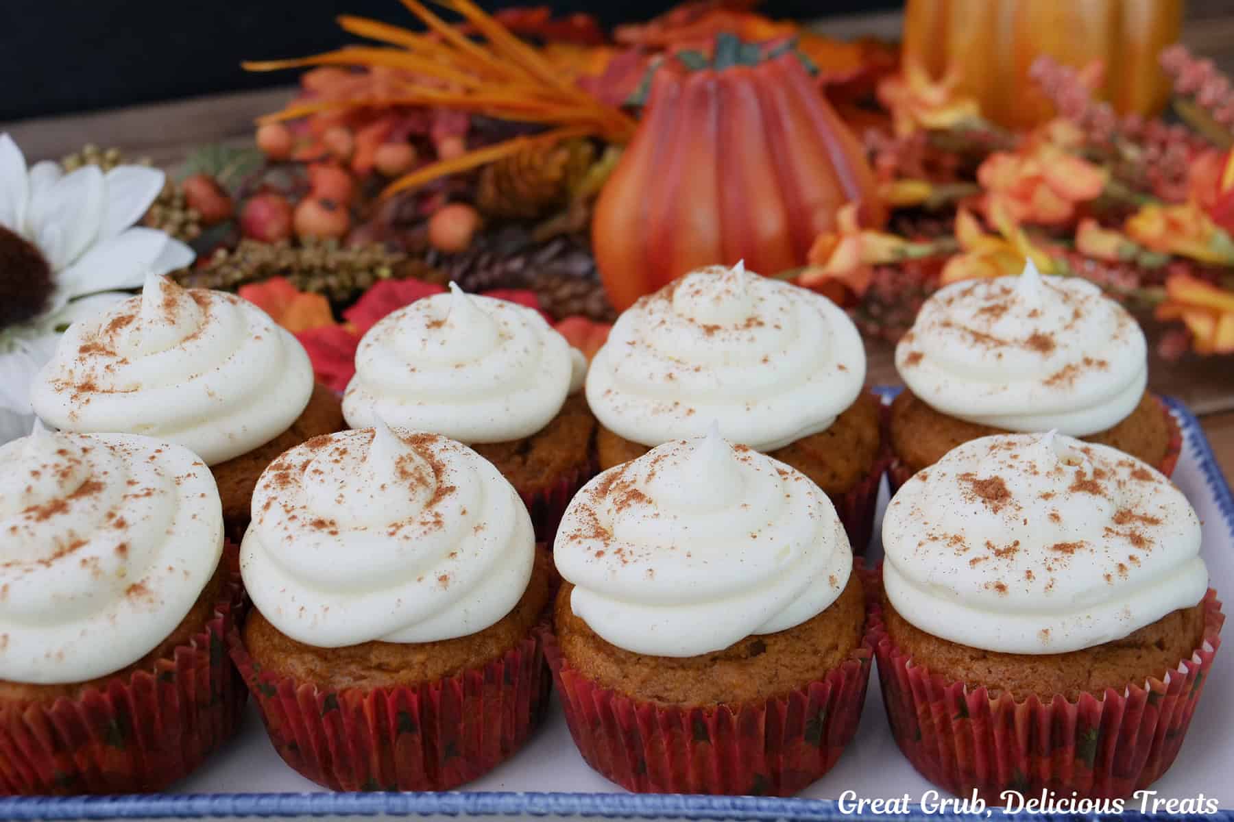 A white plate with eight pumpkin cupcakes with maple frosting on it with fall foliage in the background.