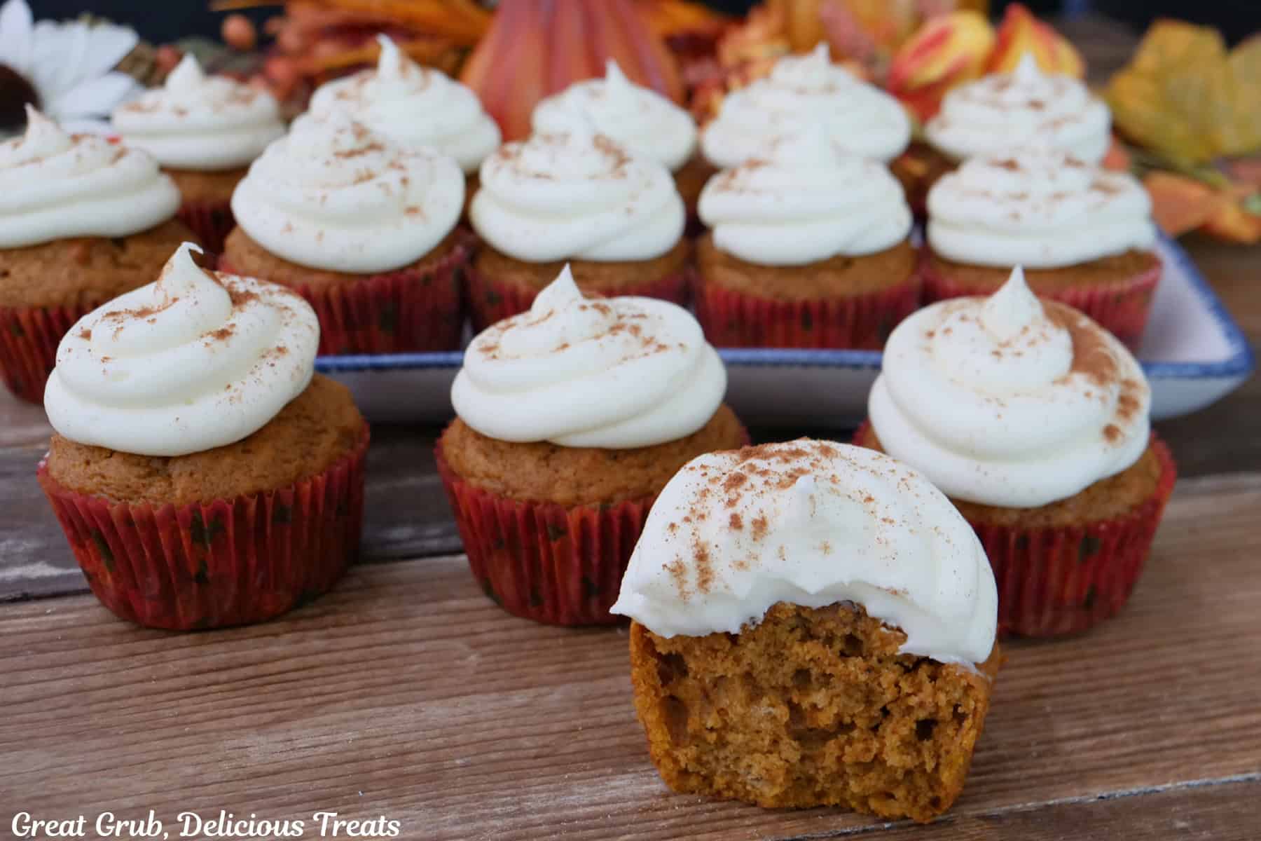 A wood surface with fourteen pumpkin cupcakes on it with a bite taken out the the cupcake in front.