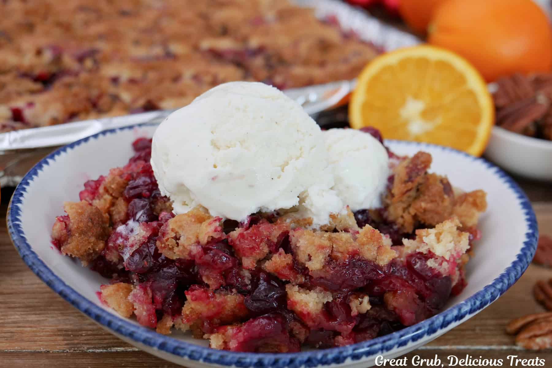 A serving of sheet pan cranberry orange cobbler with a scoop of vanilla ice cream on top in a white bowl with blue trim, and oranges and cranberries in the background.