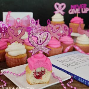 A single white cupcake with a bite taken out showing the raspberry filling in side, and more cupcakes in the background, and a book, Real Talk, Real Hope by Dr. Loren Rourke off to the right side.