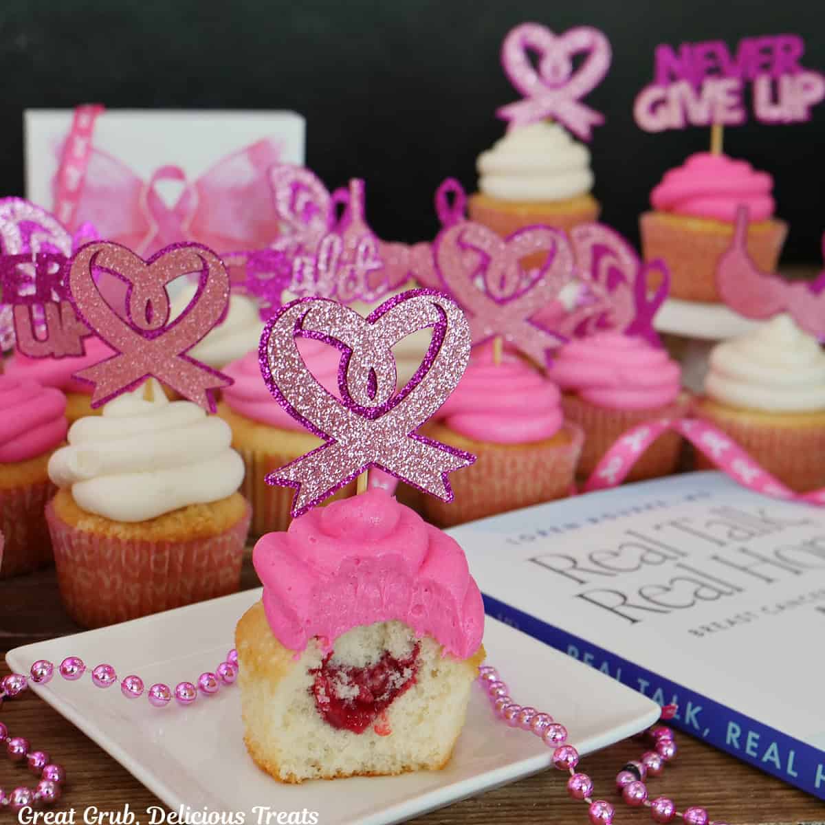 A single white cupcake with a bite taken out showing the raspberry filling in side, and more cupcakes in the background, and a book, Real Talk, Real Hope by Dr. Loren Rourke off to the right side.