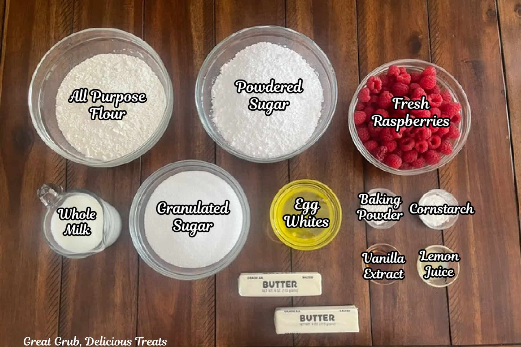 A wood surface with all the ingredients placed on it to make white cupcakes with raspberry filling and buttercream frosting.